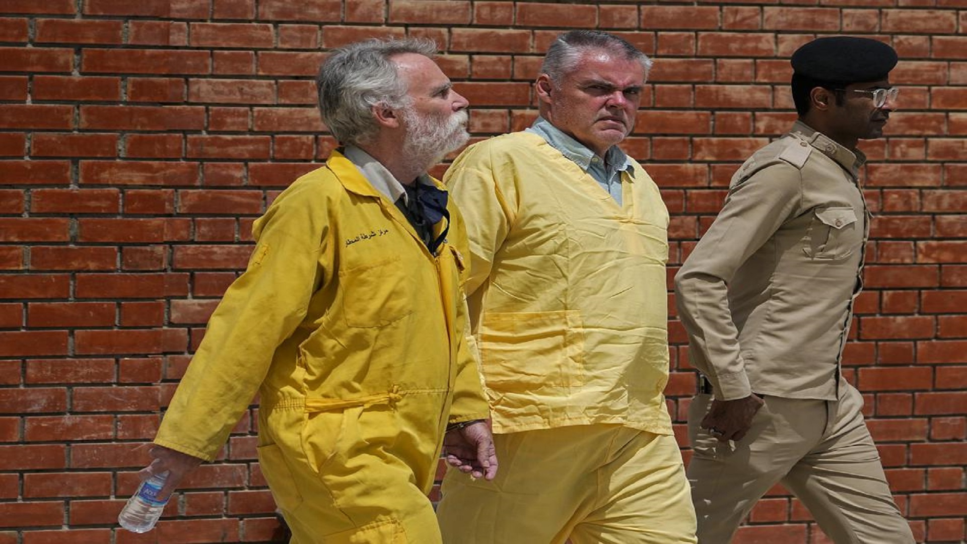 Jim Fitton of Britain, left, and Volker Waldman of Germany, center, wearing yellow detainees' uniforms and handcuffed, are escorted by Iraqi security forces, outside a courtroom, in Baghdad, Iraq, on May 22, 2022. (AP Photo/Hadi Mizban, File)